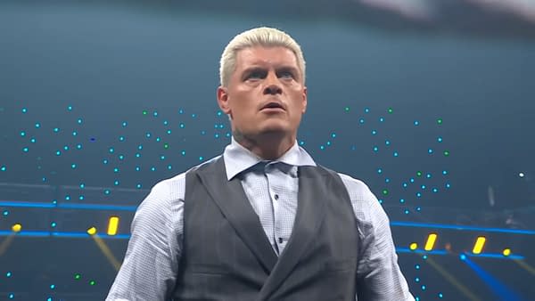 Cody Rhodes looks visibly upset during a contract signing, wearing a formal outfit with a vest and button-up shirt. The background features colorful lights, indicative of a wrestling event atmosphere.