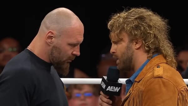 Jon Moxley and Hangman Adam Page stand face to face in a wrestling ring, both engaged in an intense exchange, with Moxley in a black shirt and Page in a brown fringed jacket.
