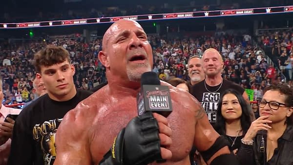 A muscular man in wrestling gear, holding a microphone, addresses a large crowd at an event. He shows signs of emotion, with sweat visible on his body, as he stands surrounded by supporters and fans.