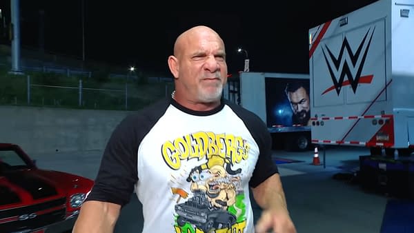 Goldberg is seen expressing determination while wearing a graphic t-shirt featuring his name and a cartoonish design. The background includes a WWE truck and a red classic car in the parking lot of WWE Raw.