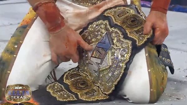 A close-up of a person's hand, belonging to Hangman Adam Page, reaching for a shiny AEW World Championship belt on the ground, adorned with jewels and intricate designs. The person's attire appears stained, suggesting an intense match.