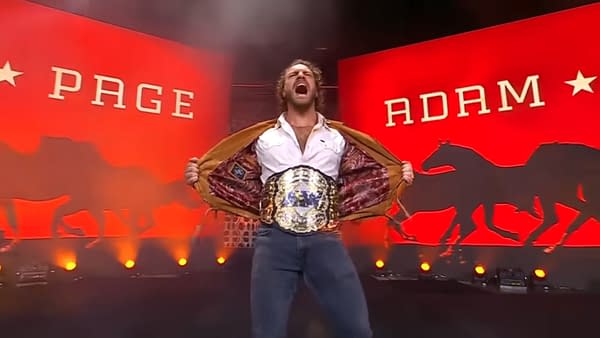Wrestler Hangman Adam Page proudly displays his newly won AEW Championship while standing on stage, holding his jacket open to display the belt. The dual screens behind page feature bright red lights and the words 'PAGE' and 'ADAM' along with silhouettes of galloping horses as Page makes his entrance on AEW Dynamite.