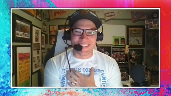 Ian Riccaboni smiles while speaking on the AEW Unrestricted podcast, wearing glasses and a headset, with a collection of framed memorabilia in the background.