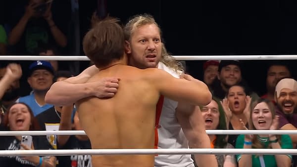 Kenny Omega and Kita Ibushi share an emotional embrace in the wrestling ring, surrounded by jubilant fans celebrating a milestone event. The two athletes display strong emotions as they momentarily set aside the intensity of competition.