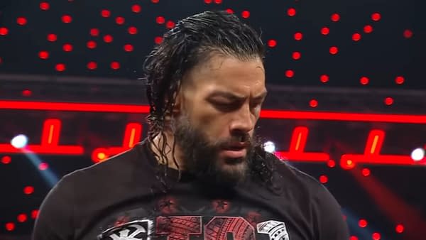 A focused Roman Reigns stands in the wrestling ring on WWE Raw, wearing a black shirt. His hair is long and wet, and there is a backdrop of bright red lights illuminating the scene.