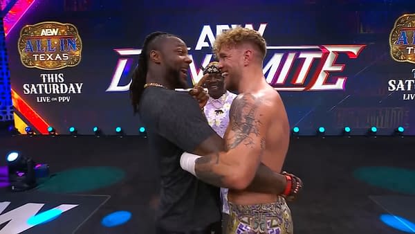 Swerve Strickland and Will Ospreay embrace in a celebratory moment on stage, with a comedic third person behind them. They are in the ring area of AEW Dynamite, featuring a colorful backdrop and bright lighting.