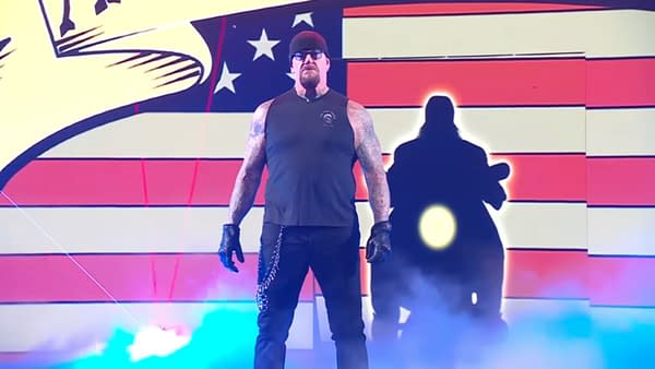 The Undertaker stands in a wrestling ring, wearing a sleeveless black shirt, black gloves, and sunglasses, with a dramatic red, white, and blue backdrop. A mist surrounds him, enhancing the theatrical atmosphere.