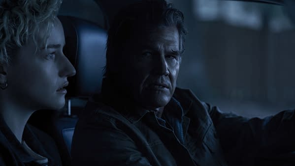 A dramatic still from the film 'Weapons' featuring two characters in a dimly lit car interior, expressing intense emotions. The scene captures a moment of tension between them.