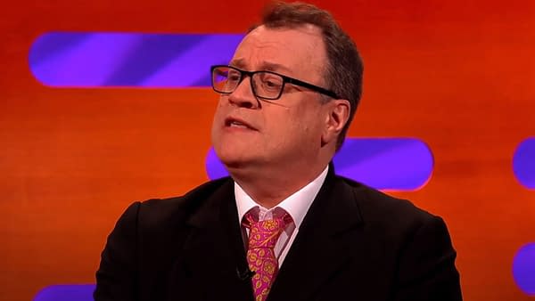 Russell T. Davies in a dark suit speaks passionately, wearing a colorful pink and orange patterned tie, against a vibrant orange and purple backdrop.