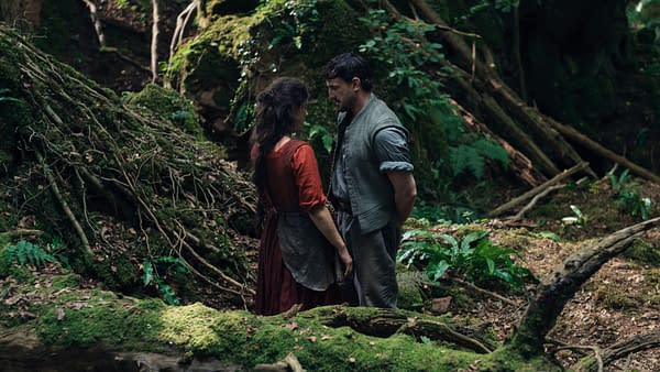 A scene from the film 'Hamnet' featuring two characters, a woman in a red dress and a man in rustic clothing, standing close together in a lush forest setting. They appear to share an intimate moment amidst moss-covered logs and greenery.