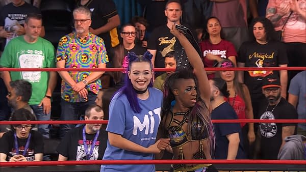 Two female wrestlers celebrate a victory in a wrestling ring. One wrestler, wearing a blue shirt, smiles broadly while the other, dressed in a colorful outfit, raises a championship belt triumphantly.