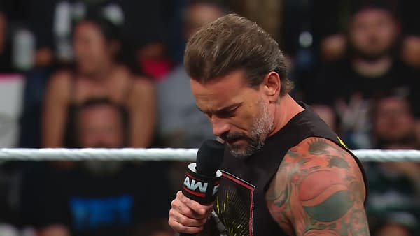 A professional wrestler, CM Punk, holds a microphone while appearing thoughtful during a segment on WWE Raw. He's wearing a sleeveless shirt and has multiple tattoos visible on his arms.