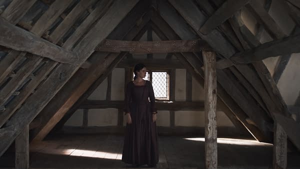 A scene from the film 'Hamnet', featuring actress Jessie Buckley as Agnes, standing alone in an attic with exposed wooden beams and a window casting light.