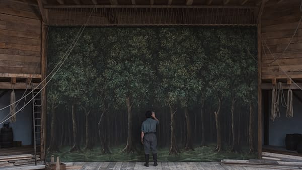 A scene from the film 'Hamnet,' featuring Paul Mescal as William Shakespeare standing in front of a large painted backdrop depicting a lush forest. The setting has wooden structures and ropes suggestive of a theater environment.