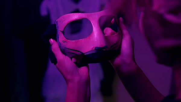 Harley Cameron holds a haunting mask with visible eye holes, surrounded by dim purple lighting, suggesting a mysterious atmosphere.