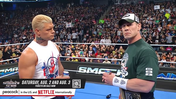 Wrestlers Cody Rhodes and John Cena stand inside the WWE SmackDown ring, sharing a beer before their match at SummerSlam. The audience can be seen in the background, cheering and holding up signs.
