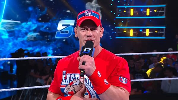 John Cena delivers a promo on WWE SmackDown while holding a microphone, wearing a bright red shirt and matching cap with the words 'U Can't See Me'. The WWE SmackDown logo is visible in the background, with an energetic crowd looking on.
