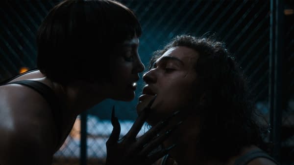 A dramatic scene from 'Kiss of the Spider Woman', featuring two characters in a close embrace, with one character's long, claw-like nails gently touching the other's face. The background is dark, enhancing the intensity of the moment.