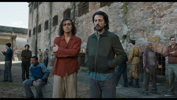 A scene from 'Kiss of the Spider Woman' featuring two main characters in an outdoor prison setting, surrounded by several other inmates. One character, dressed in a red shirt, stands with crossed arms, while the other, in a green jacket, looks pensively into the distance.