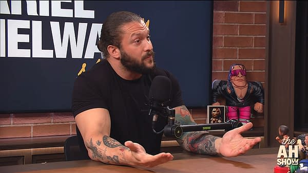 Karrion Kross speaks during a podcast interview, gesturing expressively. His tattoos are visible, and there is a wrestling figurine in the background, adding context to the setting.