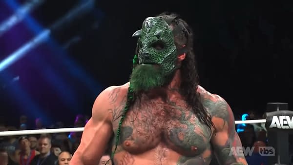 A muscular wrestler wearing a green reptilian mask with spikes and a braided hair. His upper body is heavily tattooed, and he has a green beard, standing in an arena with fans in the background.