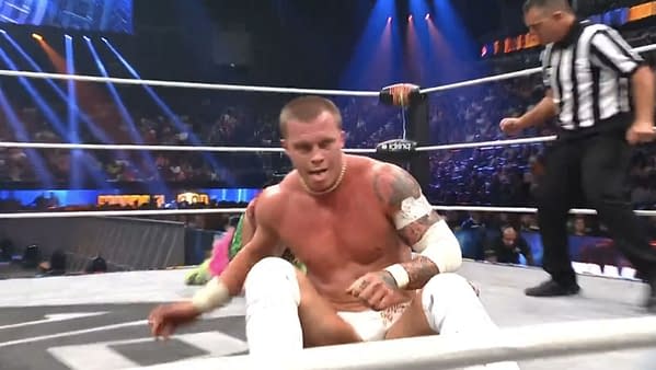 Kyle Fletcher, in wrestling gear and a determined expression, takes a moment to gloat during his match against Hiromu Takahashi. The wrestling arena is filled with lights and fans in the background.