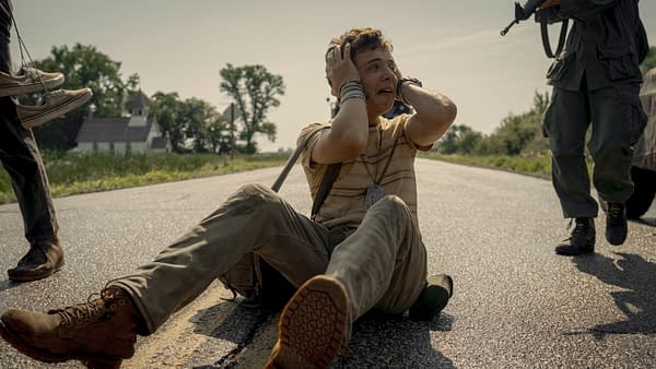 In a scene from 'The Long Walk,' a distressed young man named Curly, portrayed by Roman Griffin Davis, sits on a road with his hands on his head, expressing fear as he is approached by figures standing nearby. The setting features a rural landscape with a distant church and trees.