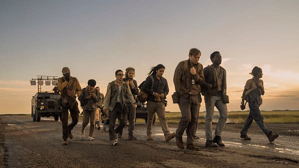 A group of young men walking along a deserted road during sunset, each carrying a bag, as a military vehicle is parked in the background. The scene depicts characters Tut Nyuot, Ben Wang, Jordan Gonzalez, Charlie Plummer, Joshua Odjick, Cooper Hoffman, and David Jonsson from 'The Long Walk'.