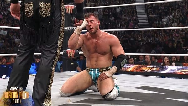 In the wrestling ring at the AEW x NJPW Forbidden Door event, MJF taunts Hangman Adam Page, who is kneeling on the mat with an expression of frustration. The crowd is visible in the background, adding to the atmosphere of the match.