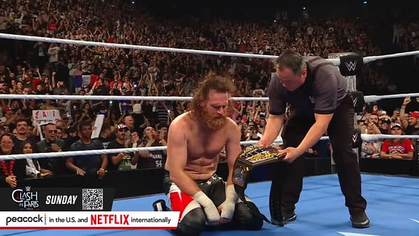 Sami Zayn kneels on the wrestling mat in celebration as a referee presents him with the WWE United States Championship after defeating Solo Sikoa in front of a roaring crowd in France.