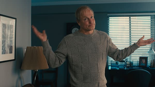 Woody Harrelson portrays Eddie McCay in a scene from the film ELLA MCCAY, showing him in a casual gray sweater with a puzzled expression, gesturing with his hands. The background includes a desk with a lamp and a photo frame.