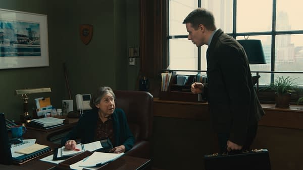 A scene from the film 'ELLA MCCAY' featuring Julie Kavner as Estelle, sitting at a desk, looking up at Jack Lowden as Ryan, who stands with a briefcase in hand. The setting appears to be an office with a green wall and a window view.