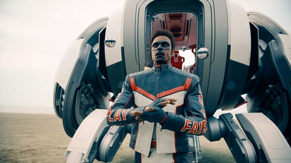 A character in a sleek, futuristic suit with red and gray accents stands in front of a large mechanical structure on a sandy beach. The character has a serious expression and performs a hand gesture.