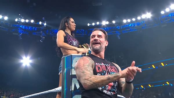 AJ Lee stands on an elevated platform, holding a championship belt and looking at the cheering crowd, while CM Punk sits beside her, clapping and smiling in response to the audience's enthusiastic reaction on WWE SmackDown.