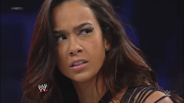 A woman with long hair, showing a concerned expression, appears in a wrestling setting. She has a serious look as she engages with the event happening around her.