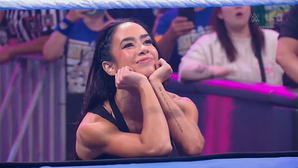 AJ Lee, a professional wrestler, sits at the edge of the ring with her chin resting on her hands, smiling warmly at the crowd during a WWE SmackDown event, with fans visible in the background. The atmosphere is vibrant and excited.