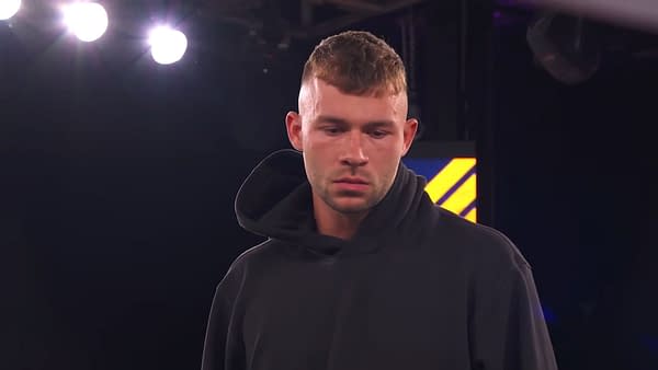 A wrestler wearing a black hoodie appears solemn as he prepares to turn heel. Bright arena lights illuminate the scene, suggesting a dramatic moment in professional wrestling.