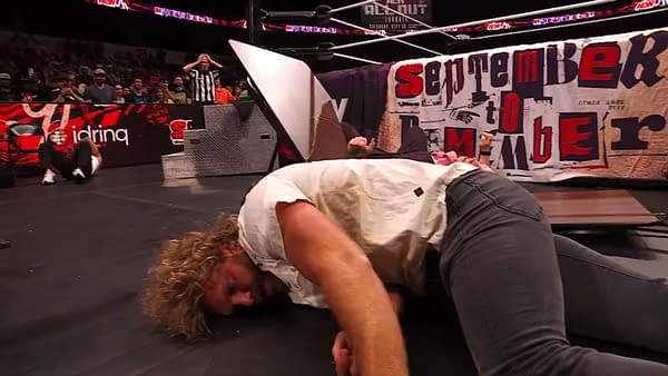Hangman Adam Page lies on the floor, visibly injured, after being suplexed through a table during an AEW wrestling event. A crowd is visible in the background, reacting to the action.