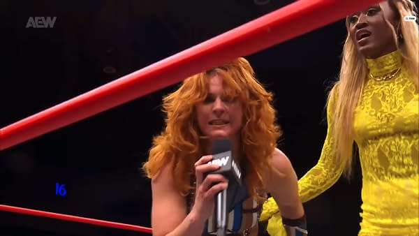 Jamie Hayter, with curly red hair and wearing a wrestling outfit, is aggressively speaking into a microphone while standing in a wrestling ring. Queen Aminata stands beside her, wearing a long yellow lace outfit and looking on.