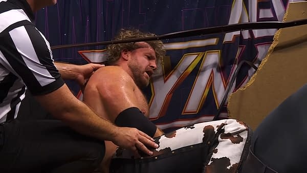 Referees tend to a hurt wrestler, Hangman Adam Page, seated near the ring after a brutal match on AEW Dynamite. The scene captures the intensity of wrestling with visible injuries and concern from the officials.