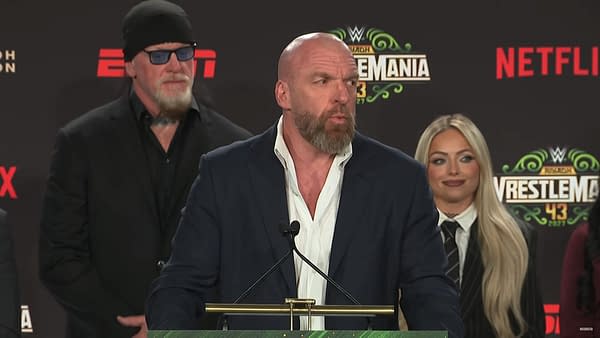 A WWE press conference featuring a well-dressed man speaking at a podium, flanked by two individuals in the background. One is in a black suit with sunglasses, while the other has long blonde hair and is wearing a striped suit.