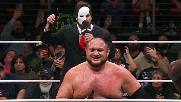 A wrestling scene features AEW Men's World Champion Hangman Adam Page, disguised as Tony Schiavone with a mask, sneaking up behind Samoa Joe, who is smiling in the ring. Fans in the background are capturing the moment with their phones.