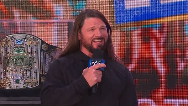 AJ Styles, wearing a black hoodie, smiles while speaking into a microphone at the Crown Jewel Perth Kickoff Show, with a championship belt displayed behind him.