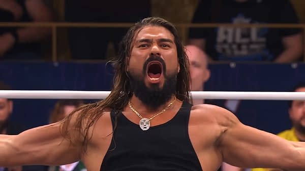 A wrestler with long hair and a beard stands in a ring, arms outstretched, passionately yelling. He wears a black tank top and a necklace, showcasing an intense expression in front of an audience.