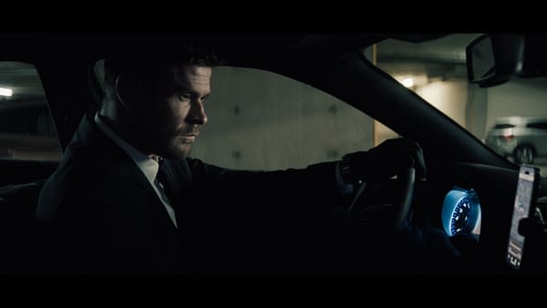A man in a black suit, portrayed by Chris Hemsworth as Davis, is sitting in the driver's seat of a car, focused on the road ahead. The interior is dimly lit, highlighting the dashboard and a smartphone displaying a navigation app.