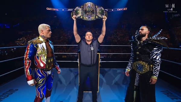 A championship belt is held high by a referee in a wrestling ring, with Cody Rhodes and Seth Rollins standing on either side, both in colorful wrestling attire. The atmosphere is electric, with an excited crowd in the background.