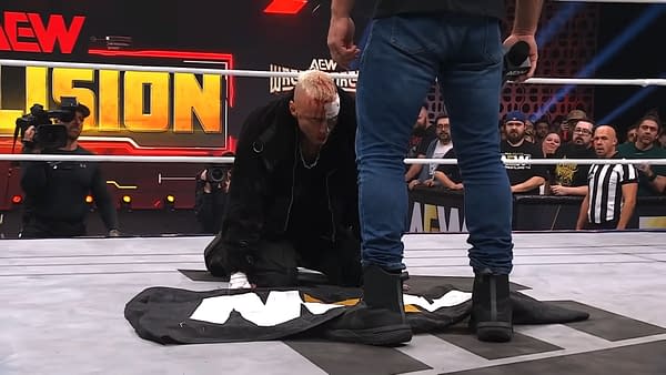 Darby Allin kneels on the wrestling ring, placing a flag on the mat as he faces Jon Moxley, who stands above him. Allin has visible injuries, adding emotional intensity to the moment.