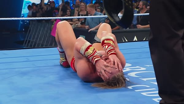 Ilja Dragunov celebrates winning the United States Championship on WWE SmackDown, lying on the mat with his hands over his face in disbelief. The audience is visible in the background, expressing excitement.
