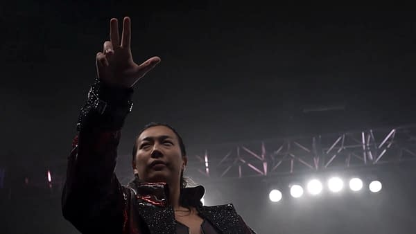 Konosuke Takeshita, dressed in a sparkling outfit, celebrates as the new IWGP Heavyweight Champion, signaling victory with a raised hand gesture in a spotlighted arena.