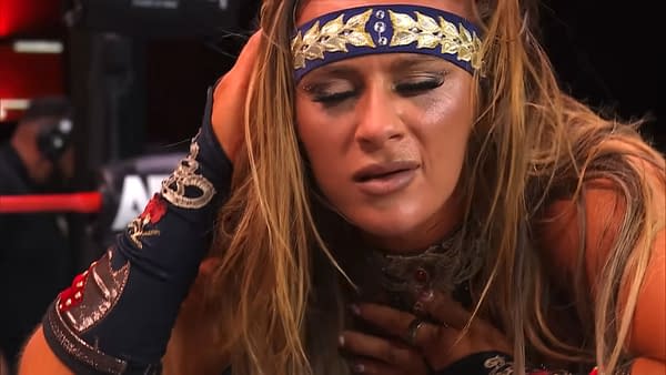 A female wrestler shows signs of distress, holding her head with a concerned expression while wearing a blue and gold headband and a decorated costume. Her makeup is slightly smudged, indicating the intensity of the moment.